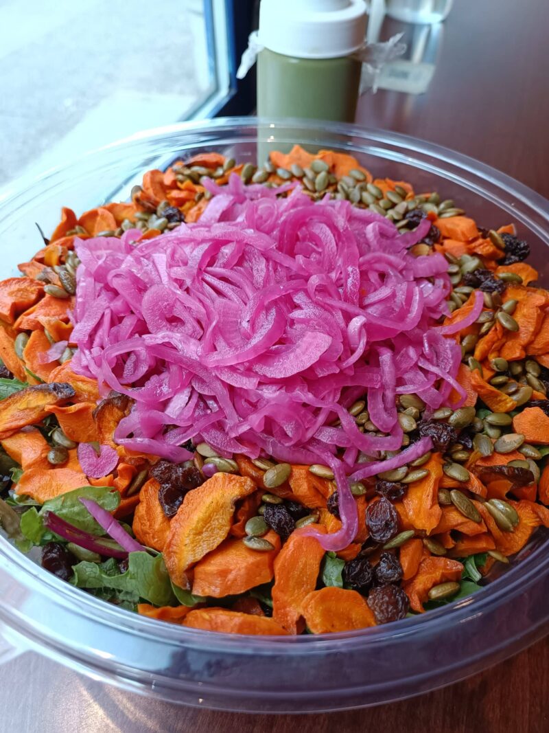 a bowl of roasted carrots salad, with hearty greens, roasted carrots, pickled red onion, raisins and toasted pumpkin seeds.