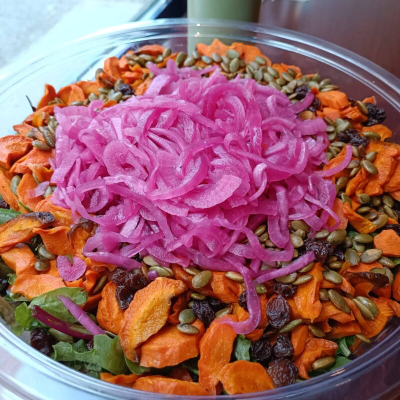 a bowl of roasted carrots salad, with hearty greens, roasted carrots, pickled red onion, raisins and toasted pumpkin seeds.