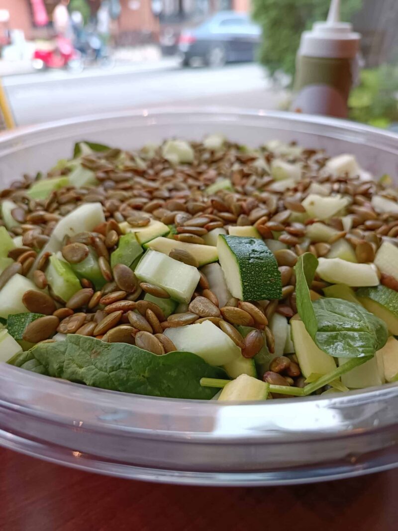 Bowl of verde salad, featuring a bed of spinach, topped with thinly sliced cucumbers, celery, zucchini and pickled green beans. Topped with crispy, spiced pumpkin seeds.