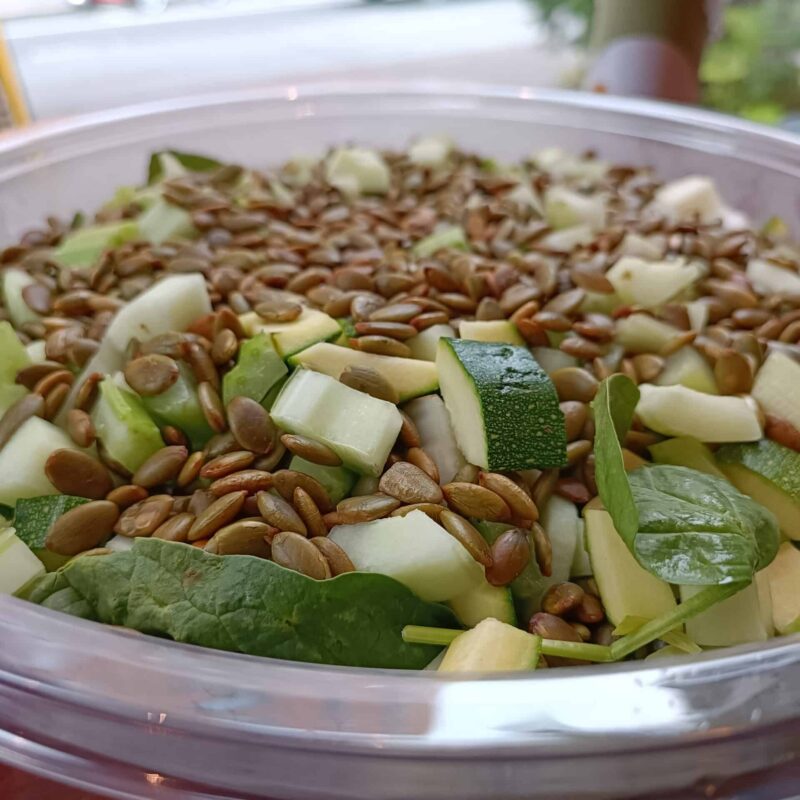 Bowl of verde salad, featuring a bed of spinach, topped with thinly sliced cucumbers, celery, zucchini and pickled green beans. Topped with crispy, spiced pumpkin seeds.